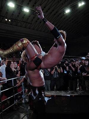 Picture of Togi Makabe holding Kazuchika Okada with a powerbomb grip before he drops him on a table outside.